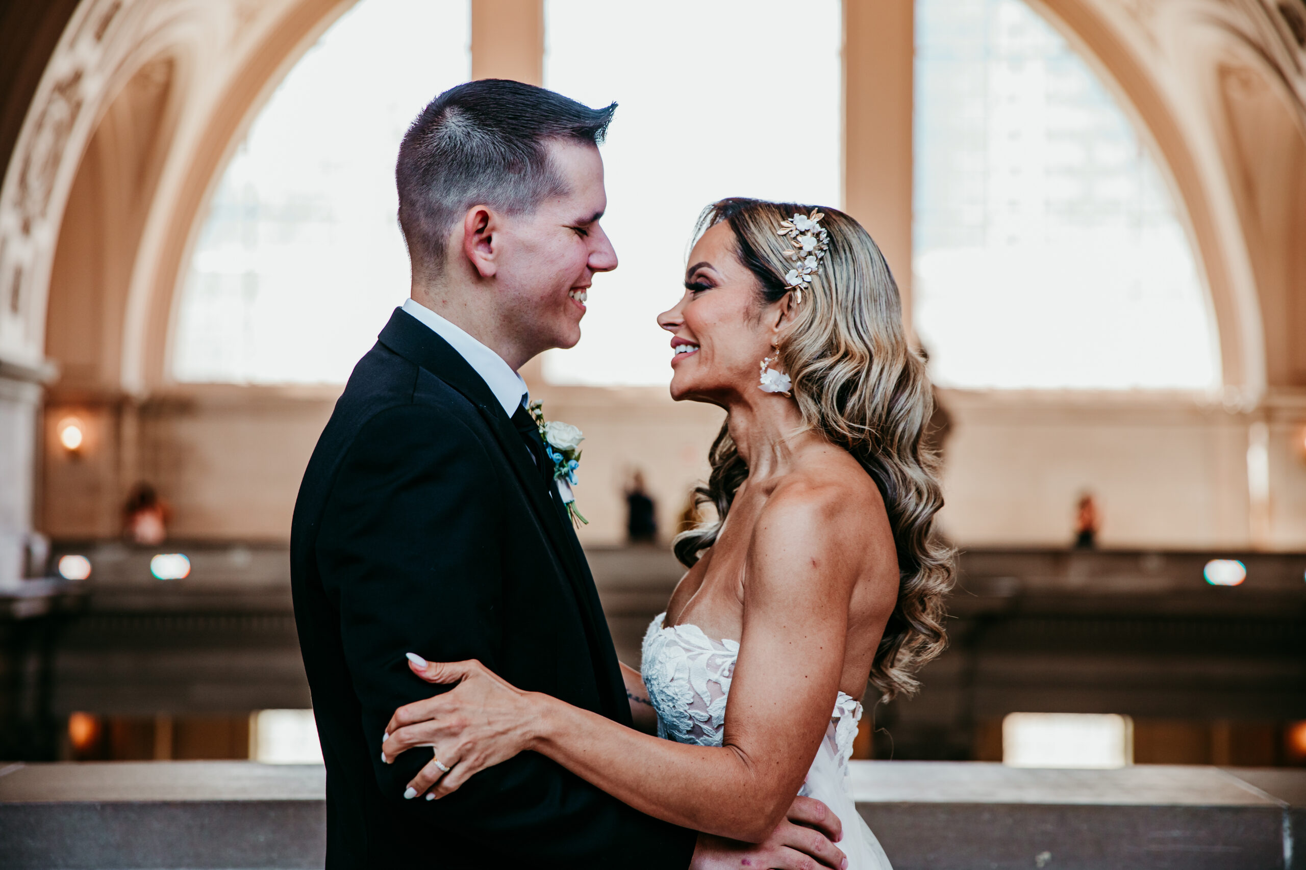 A newlywed couple shares a joyful moment inside San Francisco City Hall, gazing into each other’s eyes with smiles. The groom wears a classic black suit, while the bride stuns in a strapless lace gown with elegant accessories. The grand architecture and arched windows of City Hall provide a beautiful, softly lit backdrop.