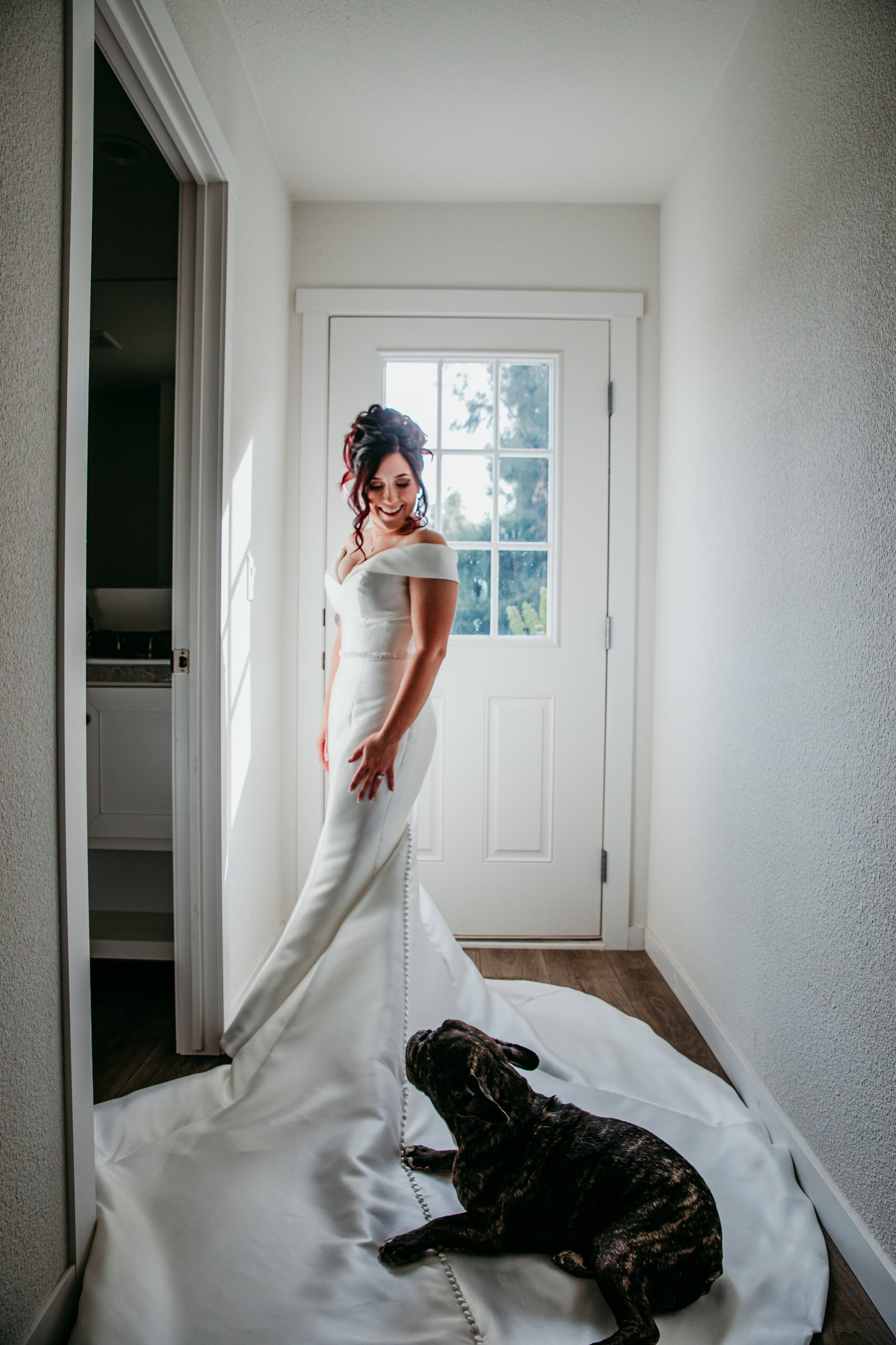 Bride in an off-the-shoulder gown smiling at her dog as it sits on her train before her fall backyard wedding in Vacaville, CA