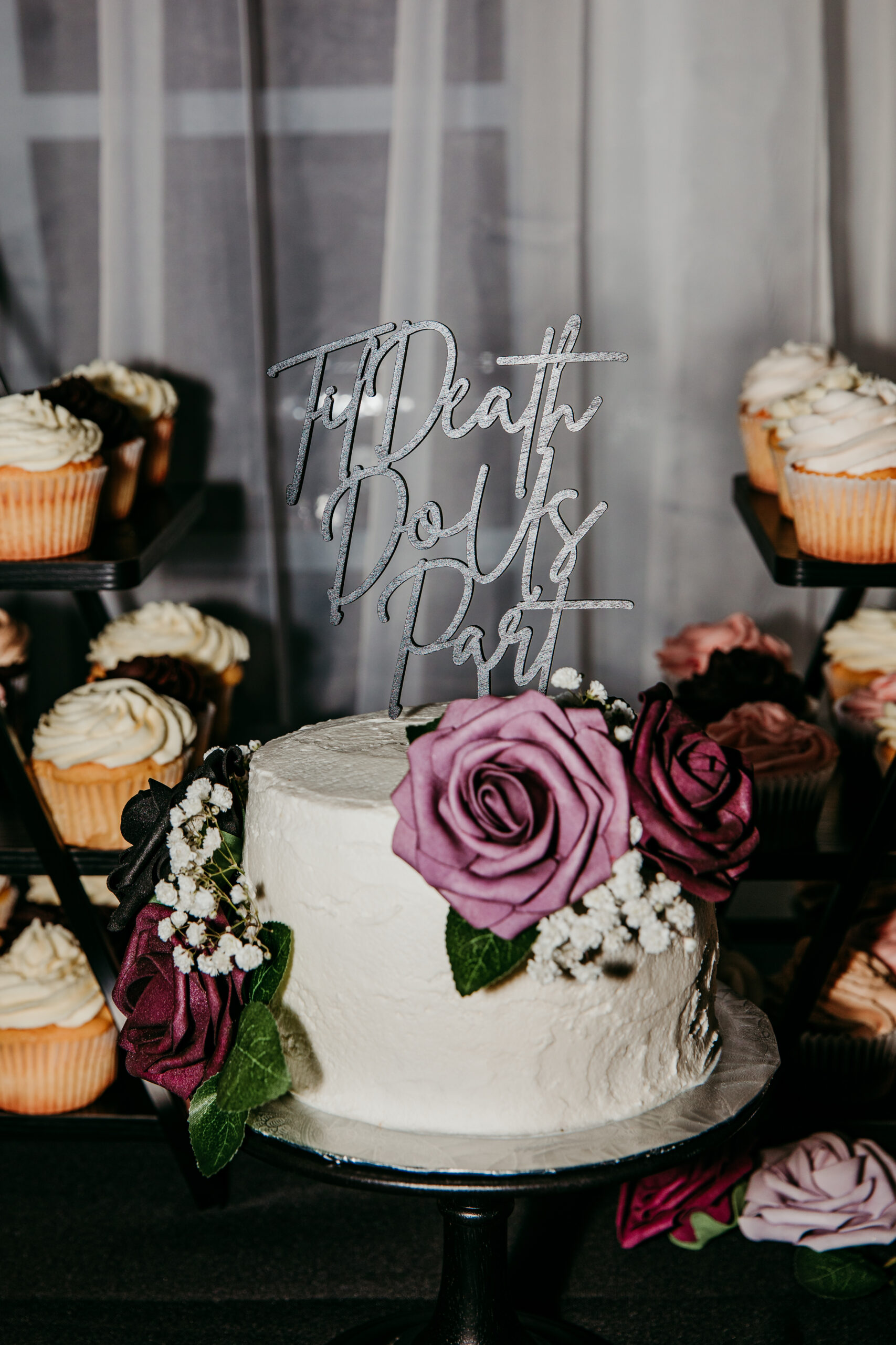 Close-up of a white wedding cake with purple rose decorations and a gothic "Til Death Do Us Part" cake topper, surrounded by cupcakes at a fall backyard wedding in Vacaville, California