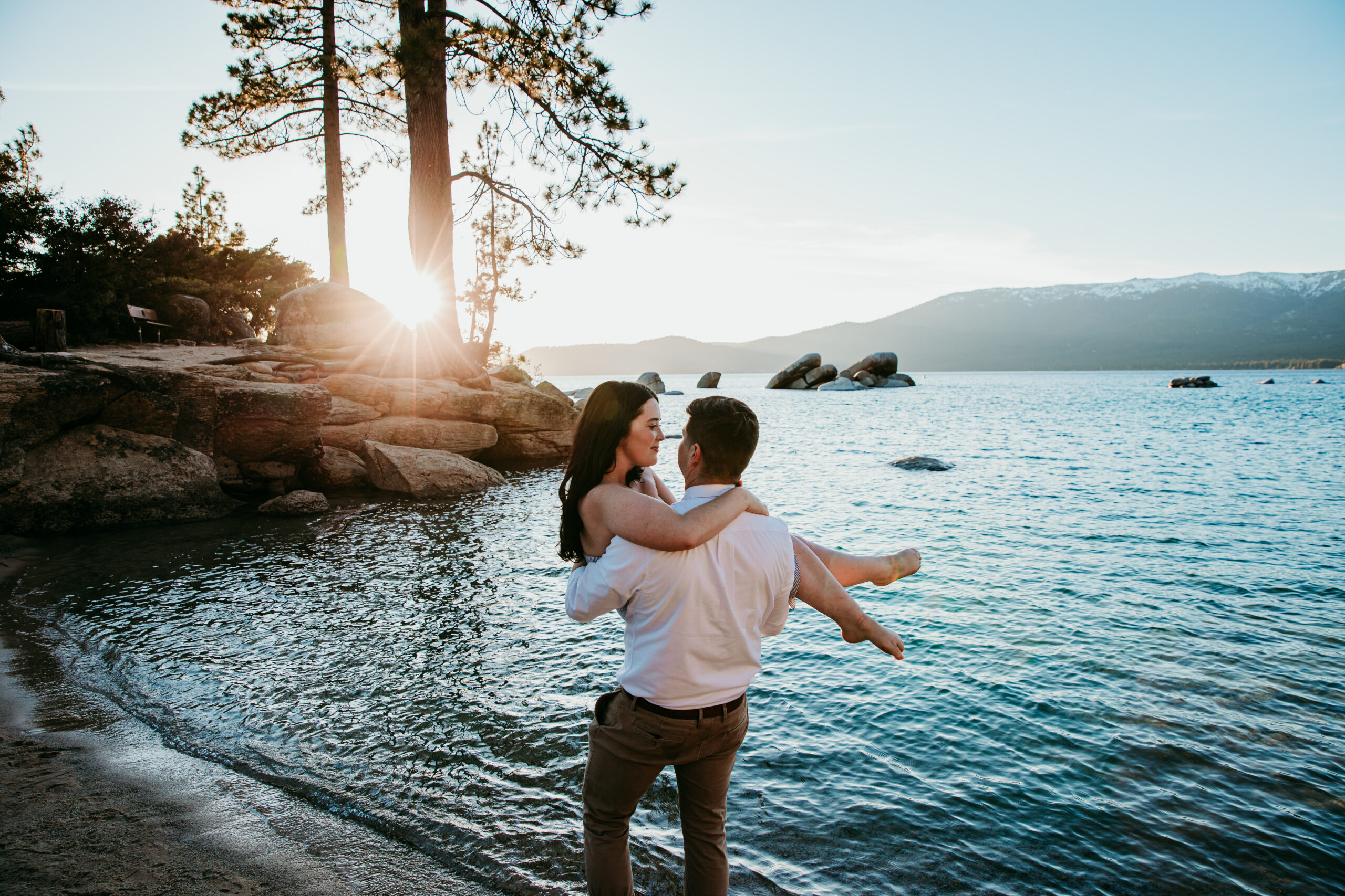 Groom lifting bride along the shoreline of Lake Tahoe at sunset, surrounded by pine trees and mountain views—an adventurous way of getting married in California.