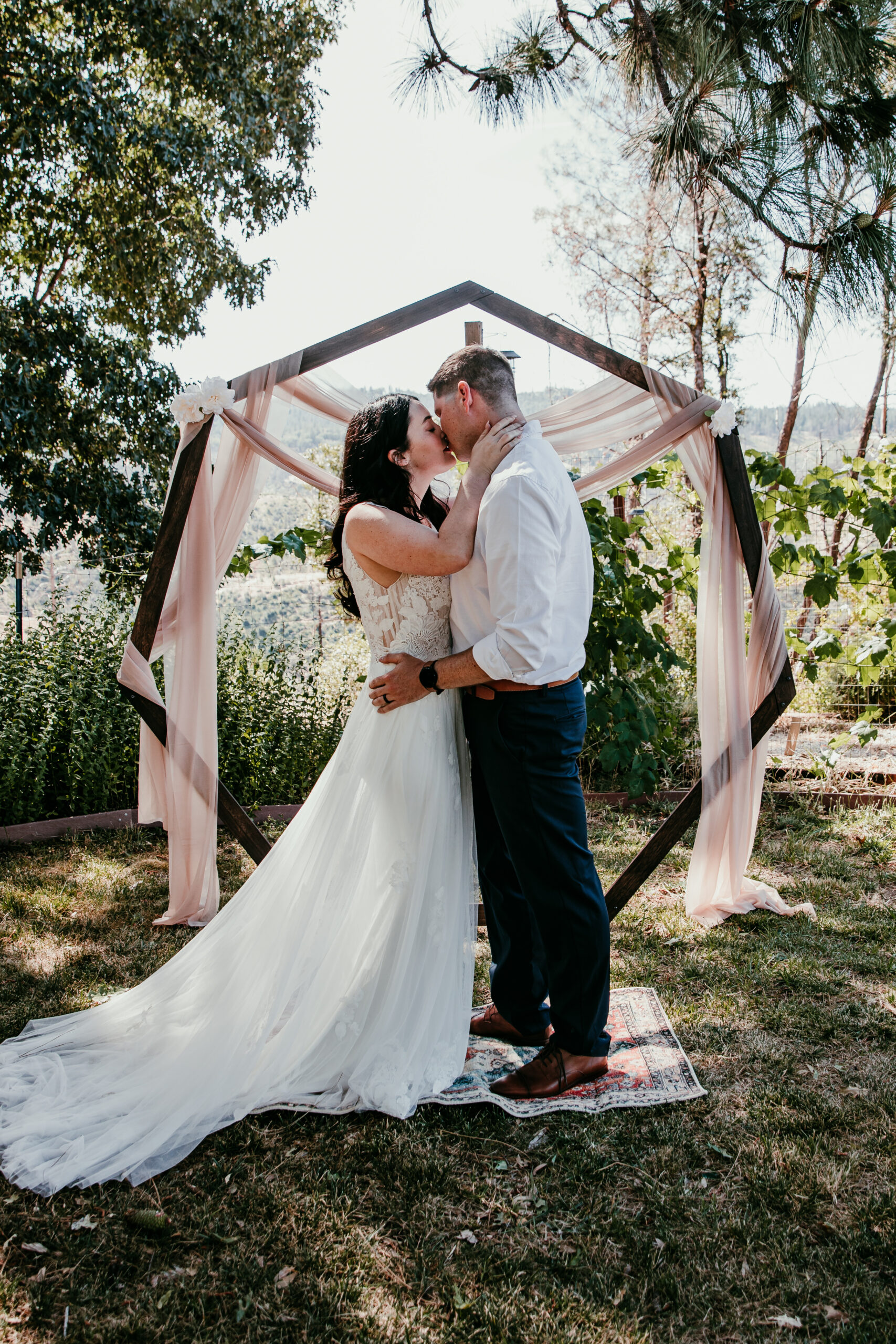 Bride and groom share their first kiss under a wooden arch during their intimate Airbnb elopement in Grass Valley CA.