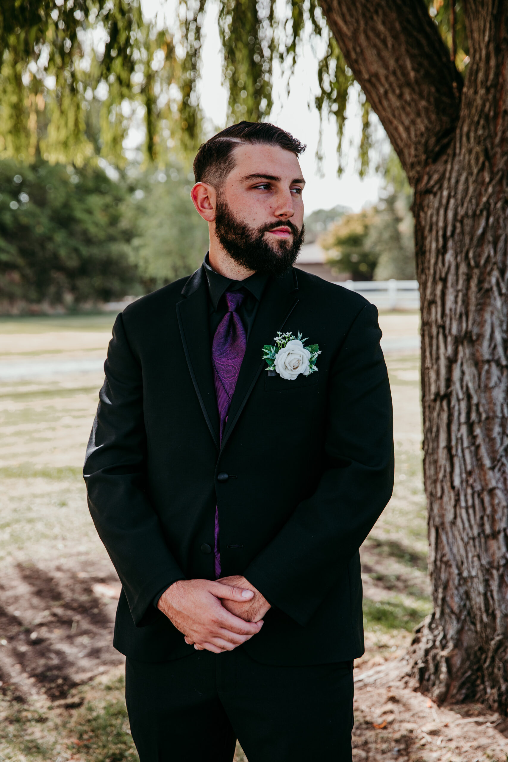 Groom in black suit with purple tie and white boutonniere standing under a willow tree before the ceremony