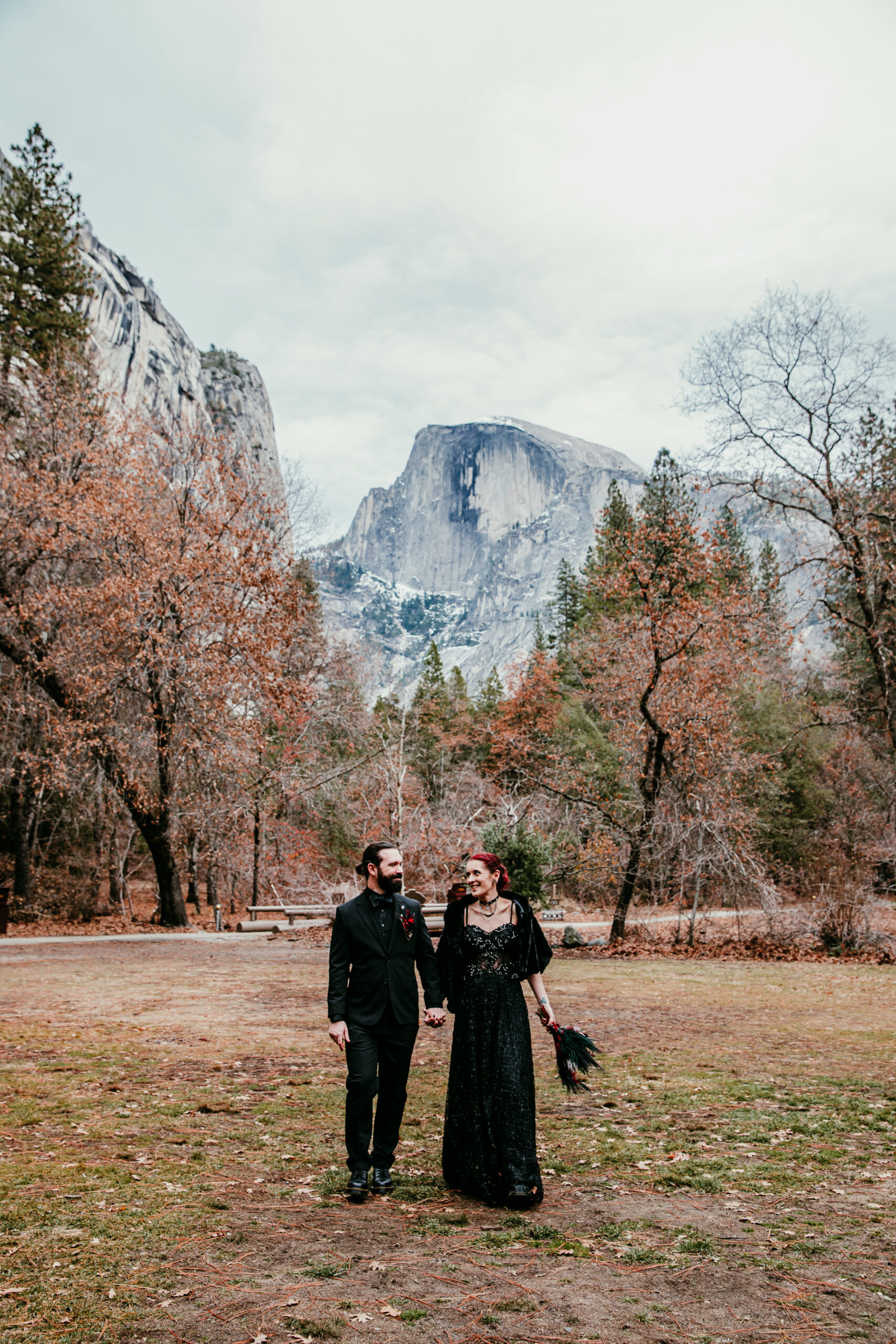 A couple walks hand in hand through Yosemite Valley during their fall elopement, surrounded by golden autumn leaves. The bride wears a black lace gown, and the groom is in an all-black suit, creating a striking contrast against the natural beauty of Half Dome towering in the background.