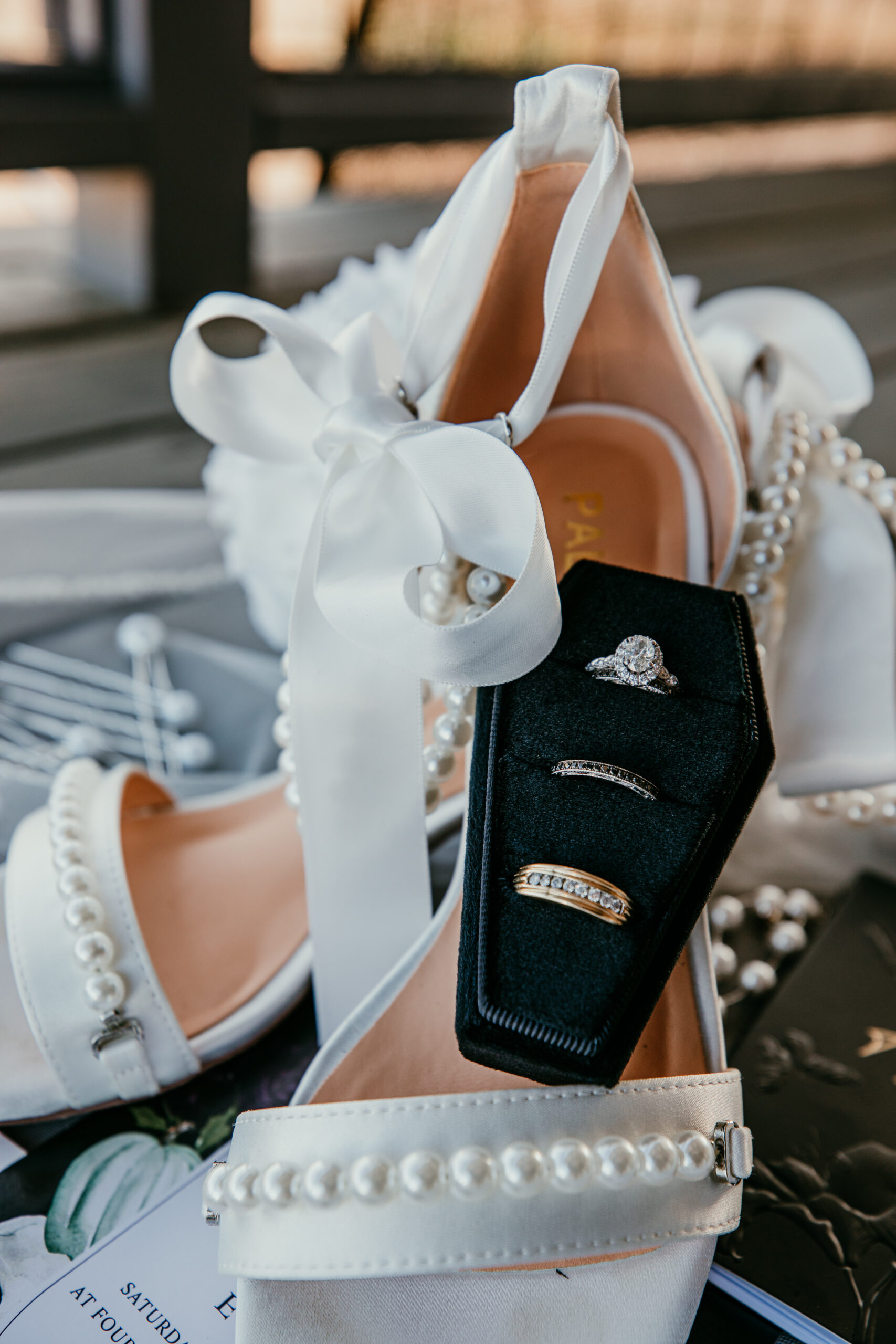 Close-up of white bridal heels with pearl straps and a black coffin-shaped ring box holding three wedding rings, styled for a fall wedding in Vacaville, CA