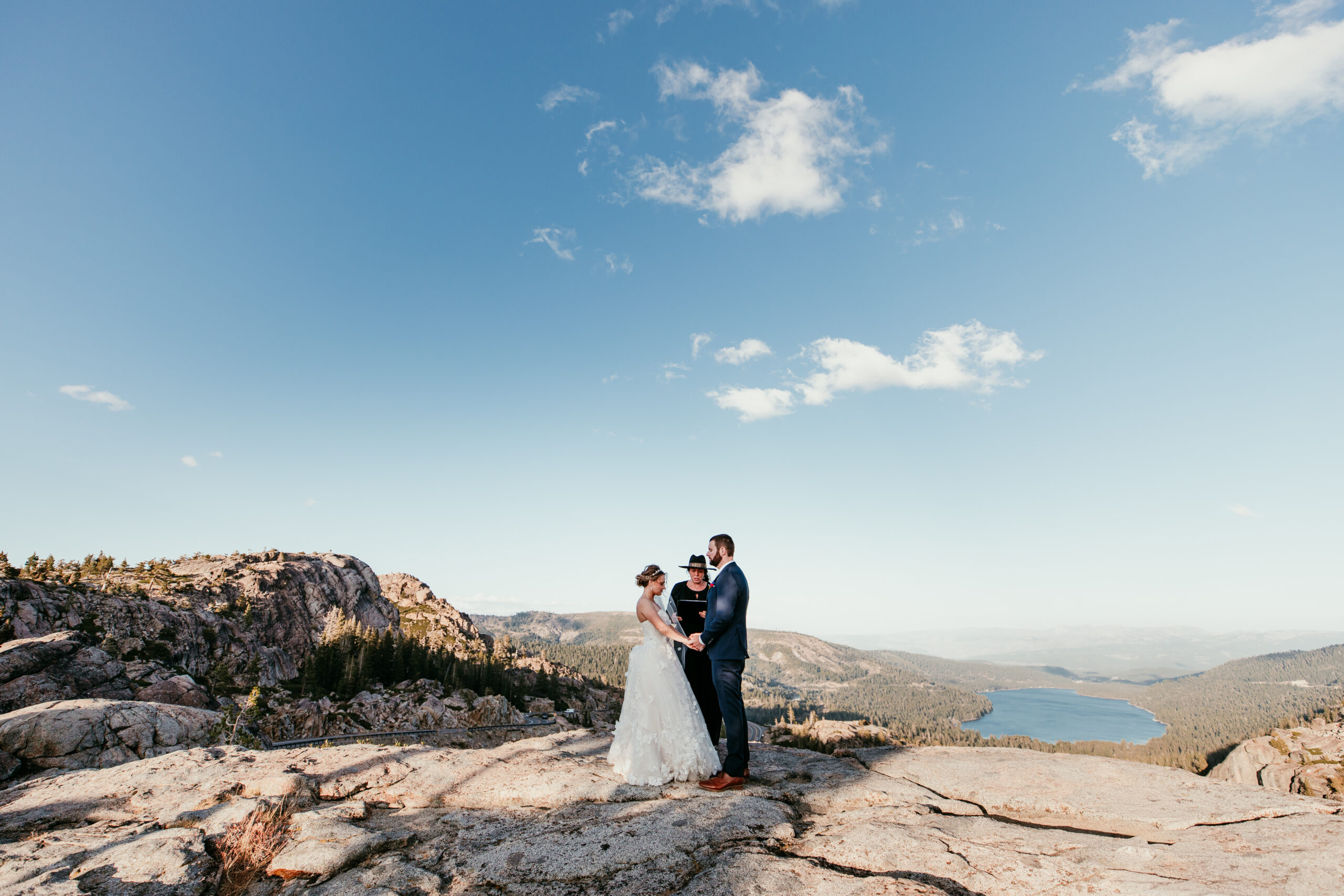 Couple holding hands with their officiant during a Donner Summit elopement ceremony, overlooking Donner Lake and forested mountains beneath a blue sky.