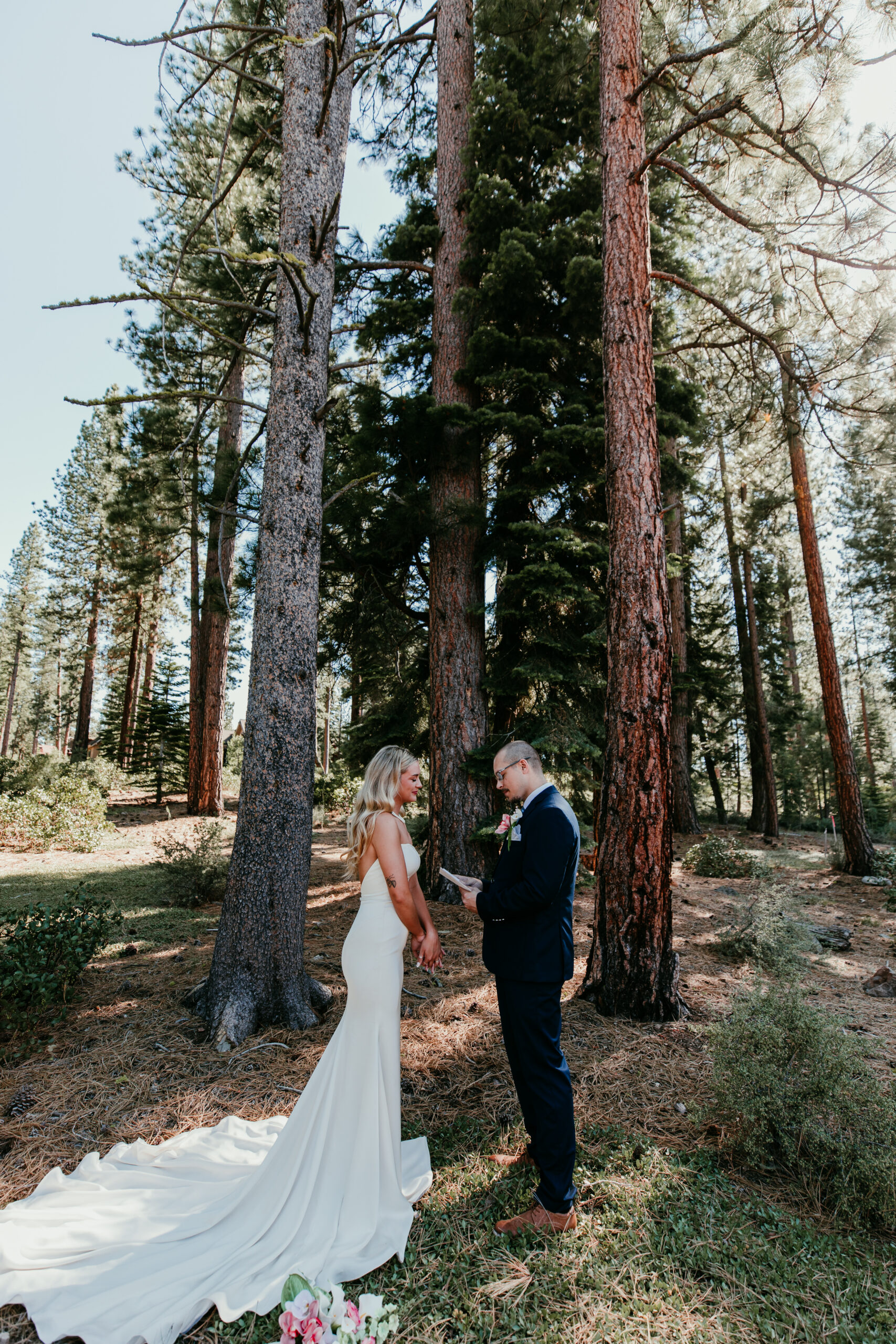 Groom reading vows to his bride during an intimate forest elopement, surrounded by tall pine trees and dappled sunlight.