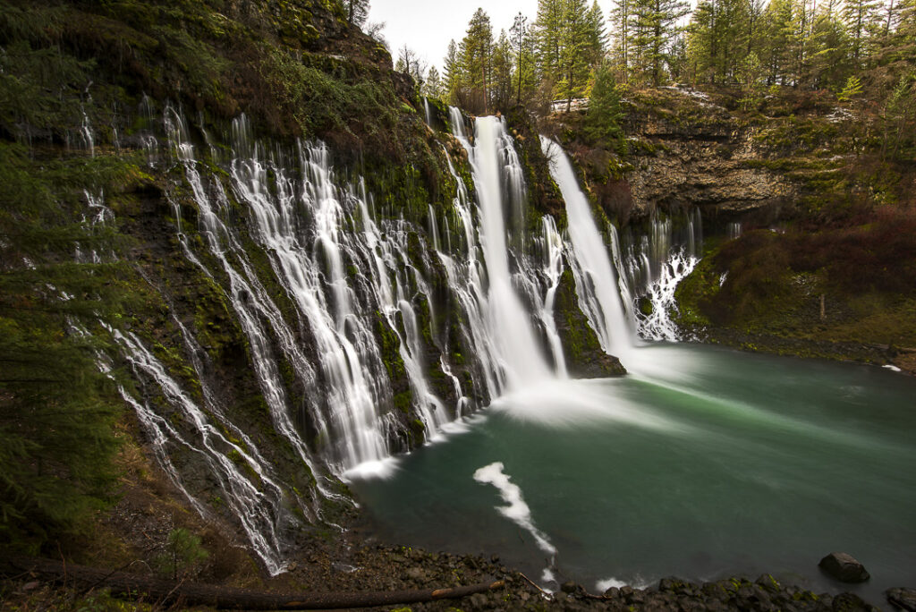 Burney Falls cascading over moss-covered volcanic cliffs into a turquoise pool, surrounded by evergreen forest in Northern California.