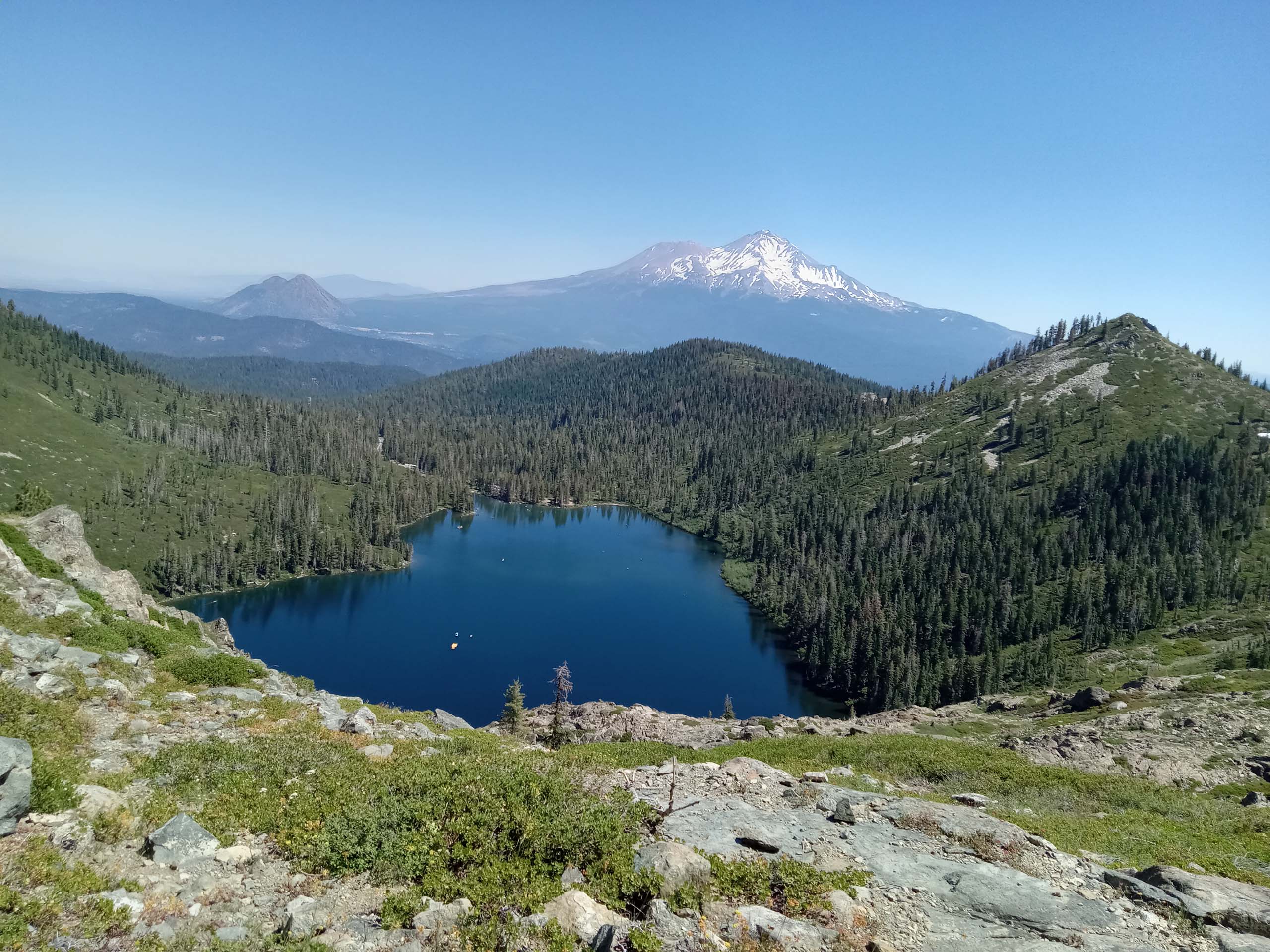 View of Heart Lake with Mount Shasta rising in the background, surrounded by alpine meadows, evergreen forest, and rocky mountain terrain.