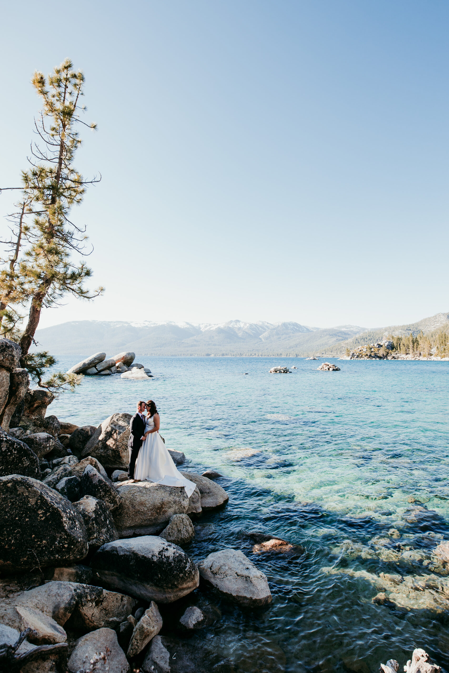 Bride and groom standing on the rocks at Sand Harbor with clear blue Lake Tahoe water and mountain views in the background.