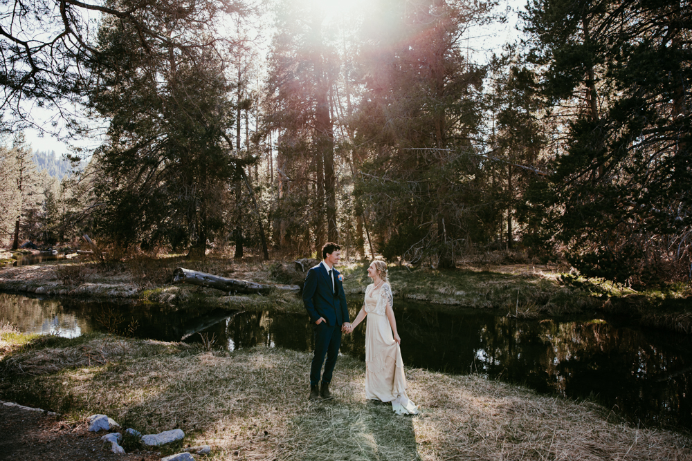 Bride and groom hold hands in a sun-drenched forest near Truckee, CA, standing beside a quiet creek surrounded by pine trees during their elopement day.