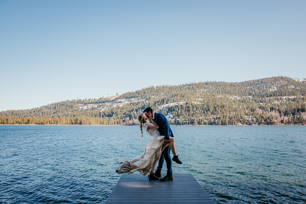 Groom lifts and kisses his bride on a dock at Donner Lake, surrounded by pine-covered mountains and sparkling blue water under a clear sky.
