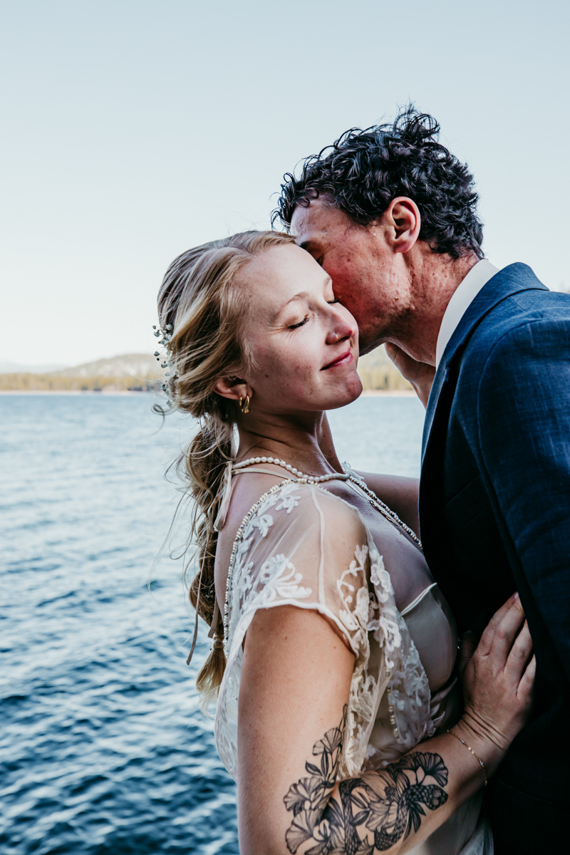 Groom softly kisses the bride’s temple as they stand close together by the water at Donner Lake, golden hour light glowing behind them.
