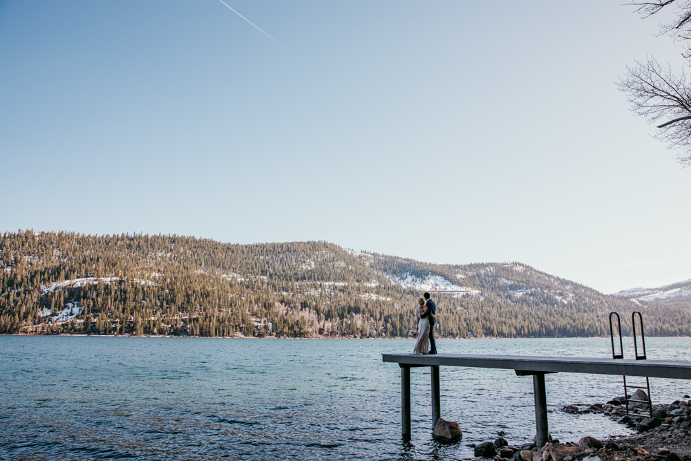 Bride and groom share a quiet moment on a dock over Donner Lake, surrounded by pine-covered mountains with patches of snow under a clear sky.