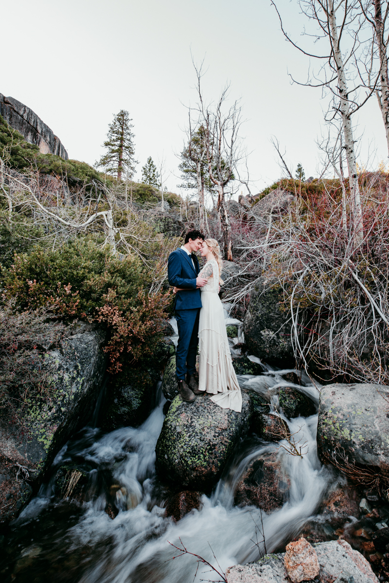 Bride and groom stand on mossy rocks above a rushing creek at Donner Summit, surrounded by early spring foliage and dramatic mountain scenery.