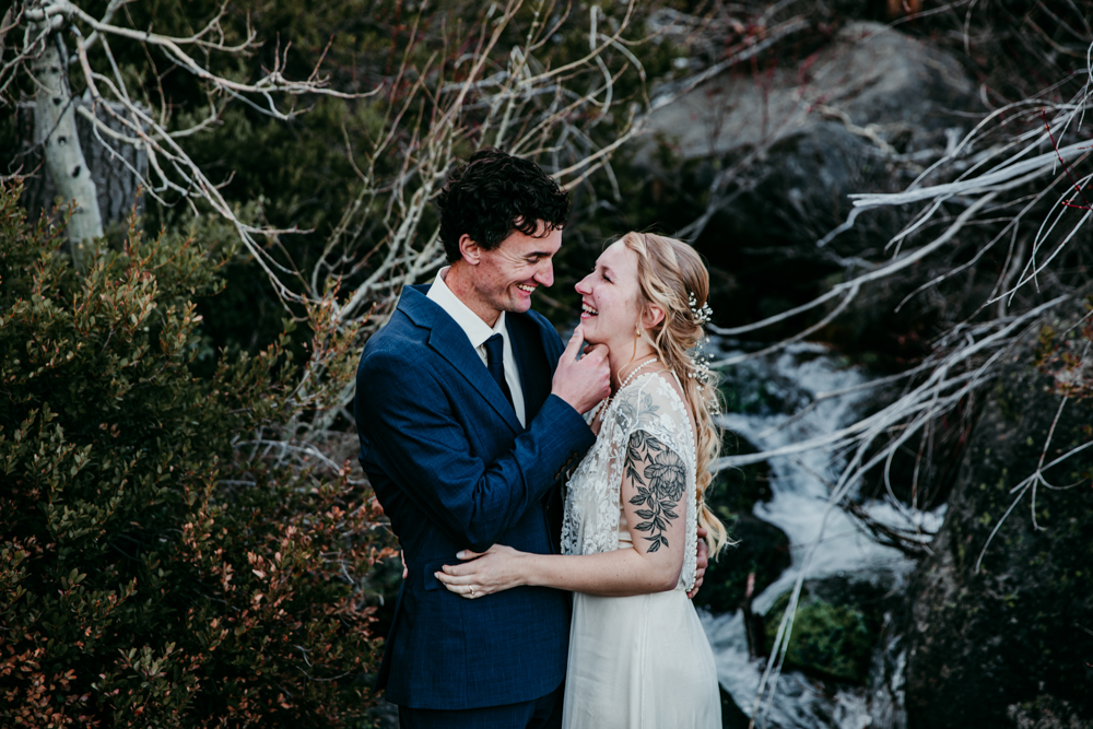 Bride and groom share a joyful moment in front of a cascading creek and spring foliage at Donner Summit during their adventurous elopement.