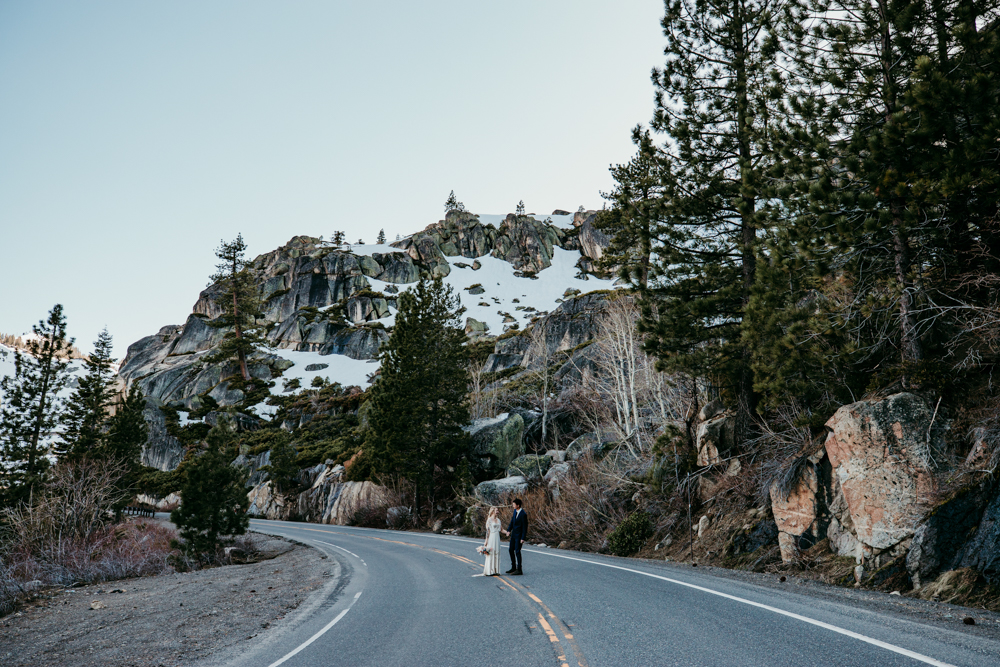 Eloping couple stands in the middle of a winding mountain road surrounded by pine trees and snowy cliffs at Donner Summit.
