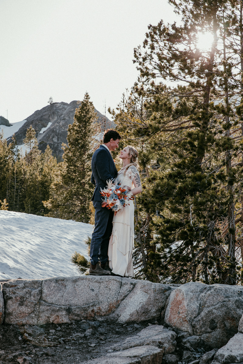 Bride and groom share a quiet moment together on a snow-covered stone ledge, surrounded by pine trees and mountain peaks at Donner Summit.