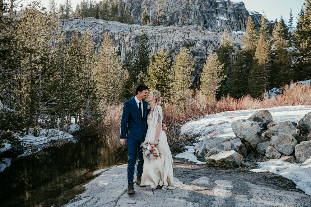 Bride and groom stand beside a creek surrounded by pine trees and snow-dusted rocks at Donner Summit, sharing a quiet, intimate moment.