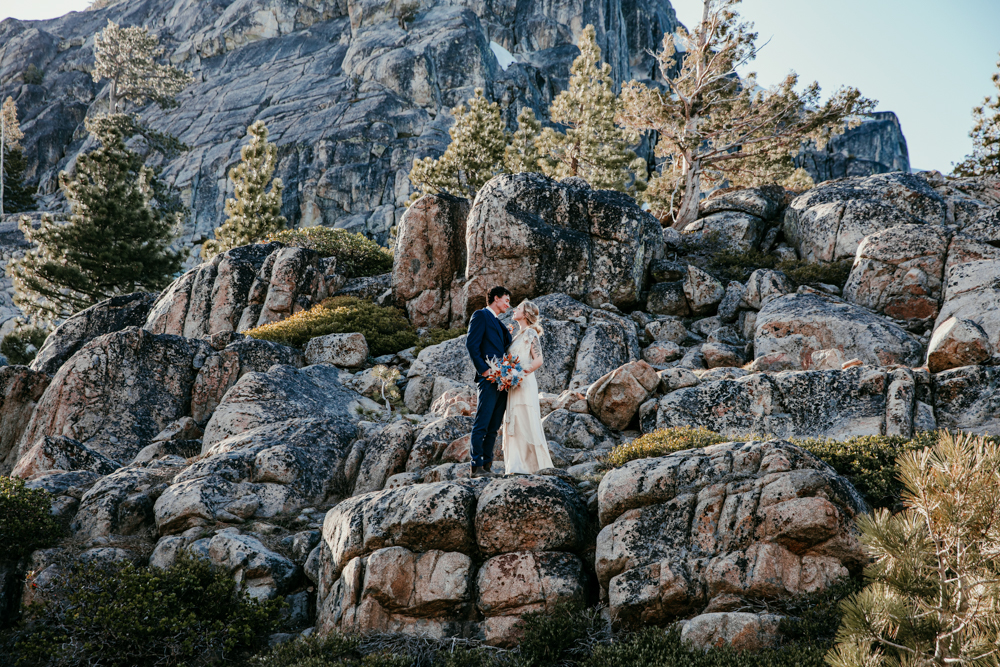 Bride and groom stand on a cluster of rugged granite boulders at Donner Summit, surrounded by towering pines and dramatic cliffside views.