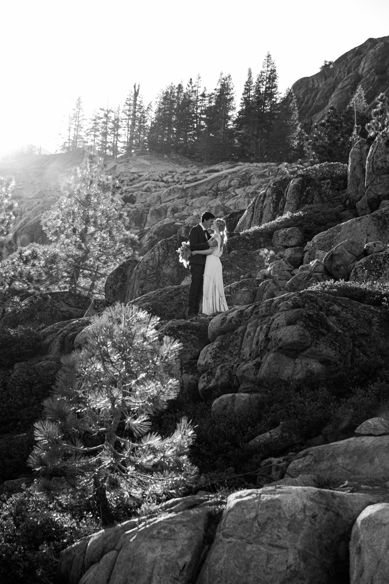 Black-and-white photo of a bride and groom embracing on rocky terrain at Donner Summit, surrounded by dramatic light and shadow with tall pines in the background.