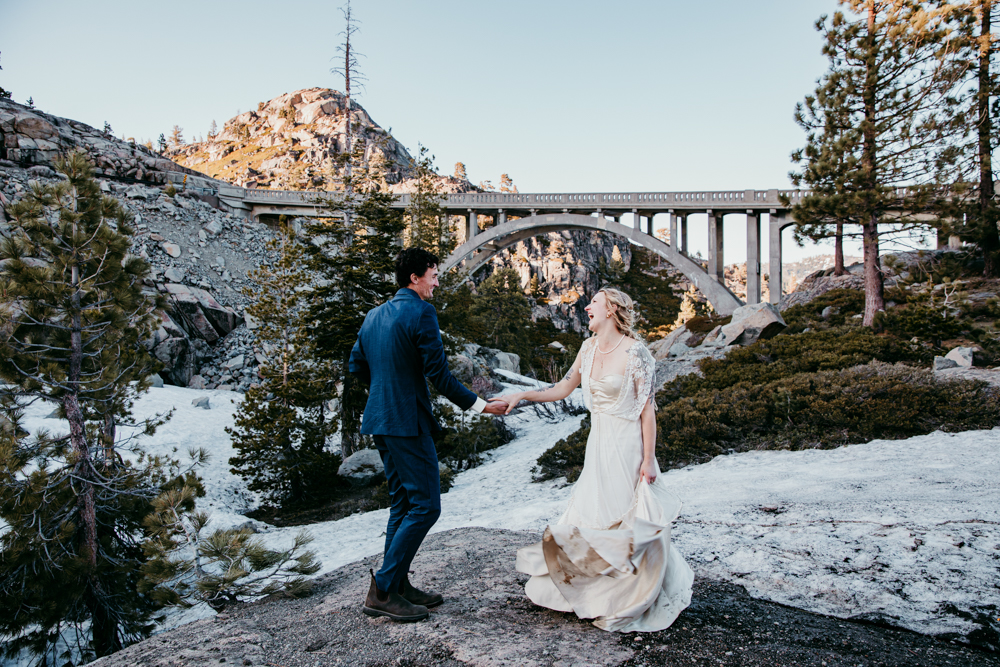 Bride and groom holding hands and walking near snow patches with the iconic Donner Summit Bridge in the background during their adventurous mountain elopement.