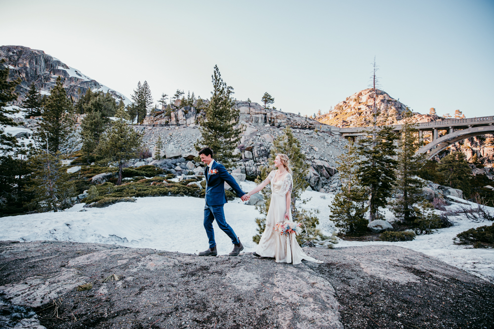 Couple walking hand-in-hand across the snowy mountain terrain at Donner Summit with Rainbow Bridge and pine-covered hills in the background.