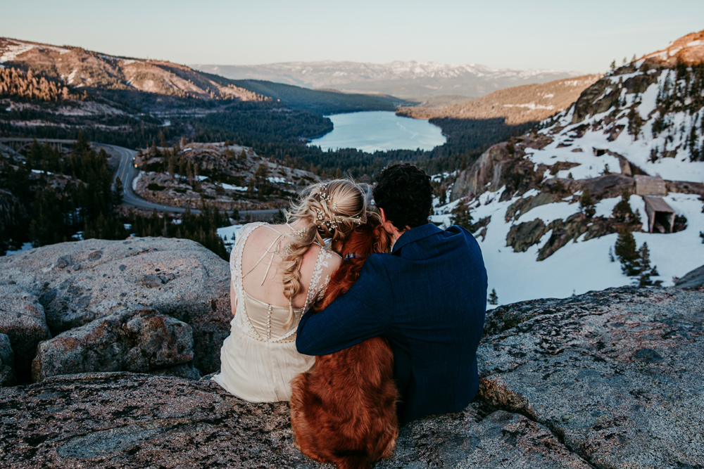Bride, groom, and their dog sitting on a rocky ledge overlooking Donner Lake at sunset, wrapped in a quiet moment together.