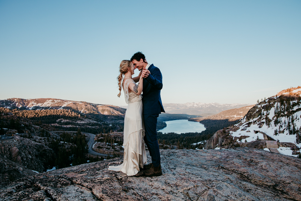 Bride and groom embracing on a rocky overlook at Donner Summit with sweeping views of Donner Lake and the Sierra Nevada mountains in the background.