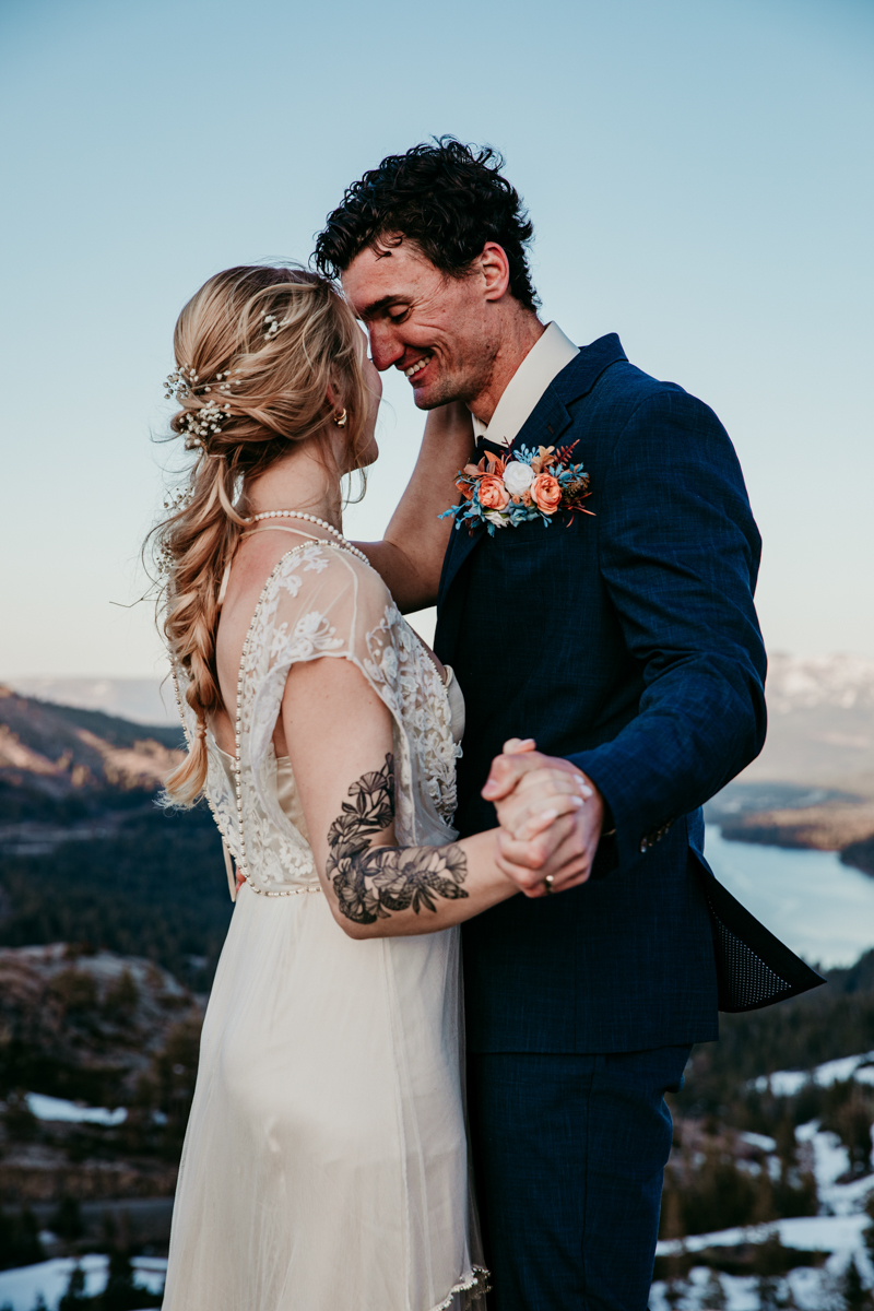 Close-up of a bride and groom smiling at each other while holding hands during their Donner Summit elopement, with scenic mountain views in the background.