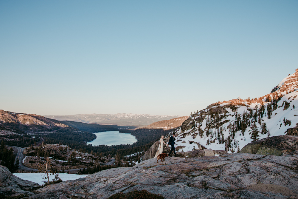 Wide scenic shot of a couple walking hand-in-hand with their dog across a rocky overlook at Donner Summit, with snow-dusted mountains and Donner Lake in the background during golden hour.