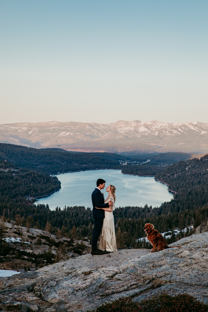 Elopement couple embracing on a mountaintop overlook with their golden retriever sitting nearby, overlooking Donner Lake and a vast forested valley at sunset.