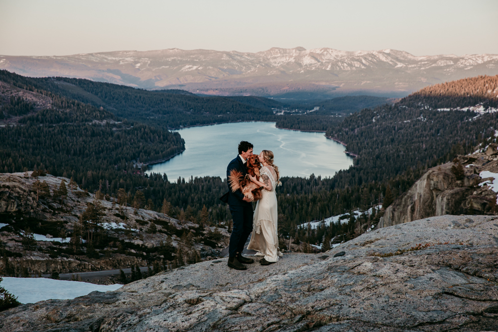 Bride and groom standing on a rocky overlook at Donner Summit, cuddling their golden retriever with Donner Lake and pine-covered mountains stretching into the distance.
