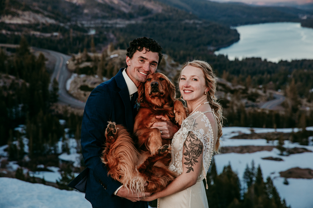 Bride and groom smiling at the camera while holding their golden retriever, standing on a snowy cliffside with Donner Lake winding through the forest below.