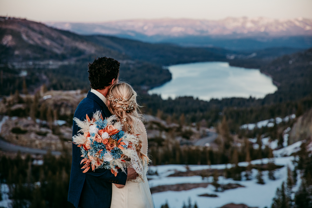 Bride and groom embrace while looking out over Donner Lake at sunset, with the bride holding a vibrant bouquet of orange and blue flowers.
