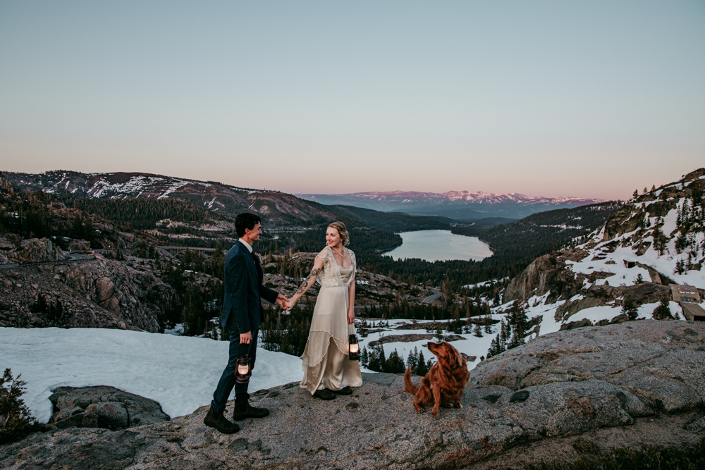 Bride and groom holding hands at sunset with their dog nearby, standing on a rocky overlook above Donner Lake surrounded by snow-dusted mountains.