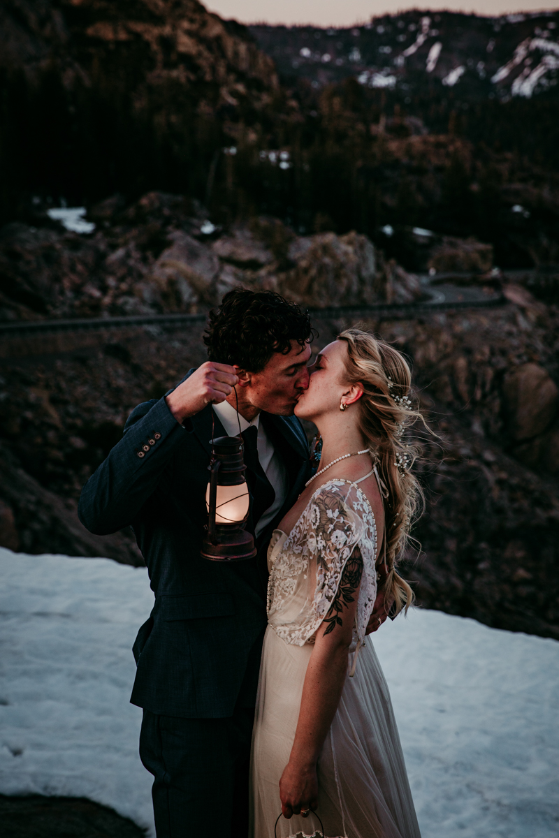 Bride and groom sharing a kiss while holding a glowing lantern, standing on snow with rugged mountain terrain in the background.