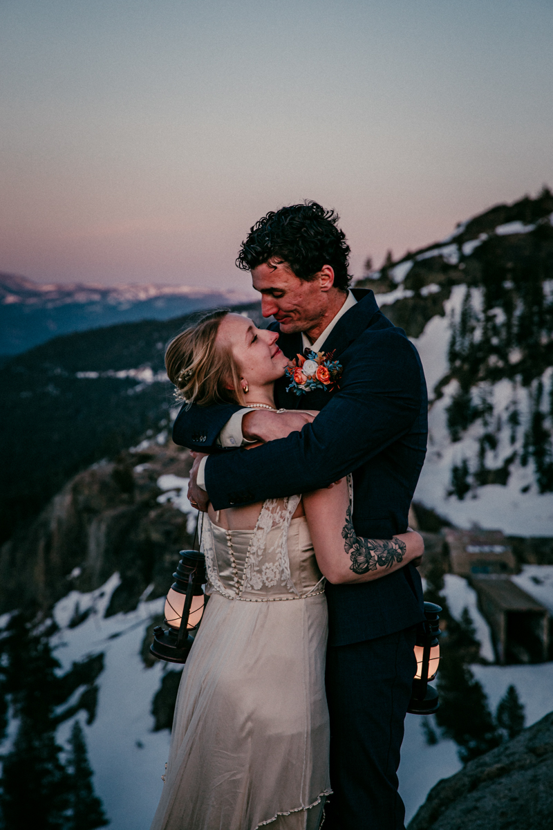 Bride and groom sharing an emotional embrace at sunset on a snowy mountainside, both holding lanterns with soft mountain light in the distance.