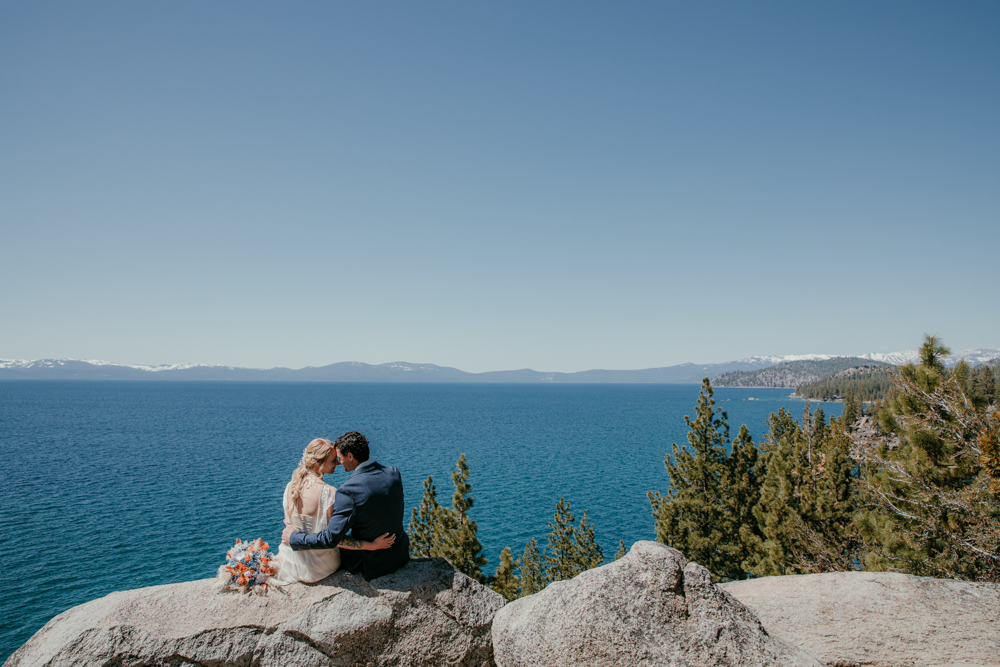 Bride and groom sitting on a rock, holding each other and looking out over the deep blue water of Lake Tahoe at their Logan Shoals Vista Point elopement