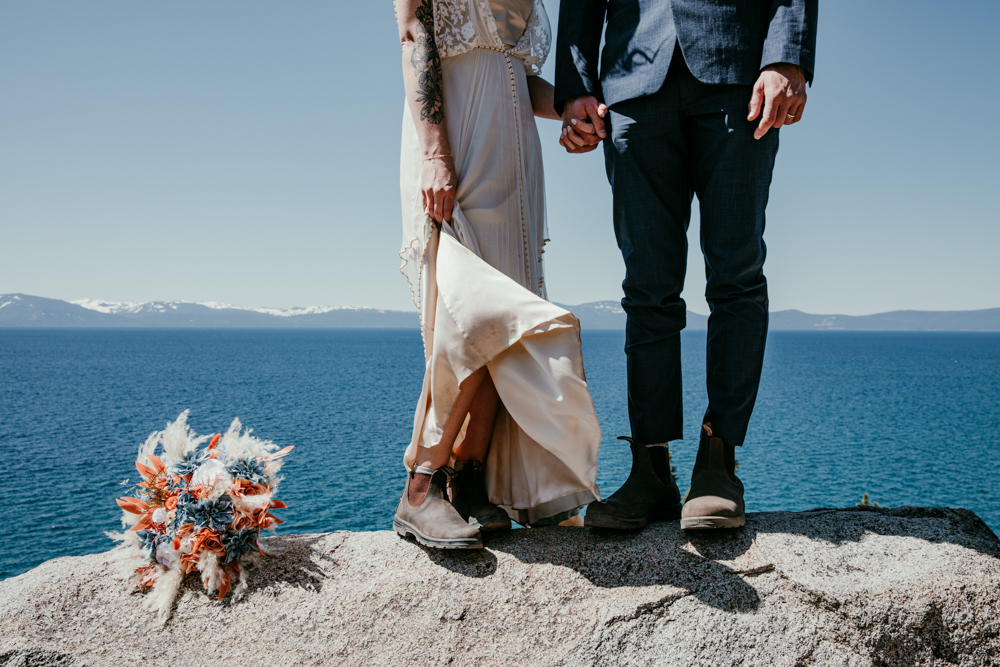 Close-up of couple holding hands on a rocky overlook at Lake Tahoe, with the bride lifting her dress to reveal Blundstone boots and a colorful bouquet resting beside them