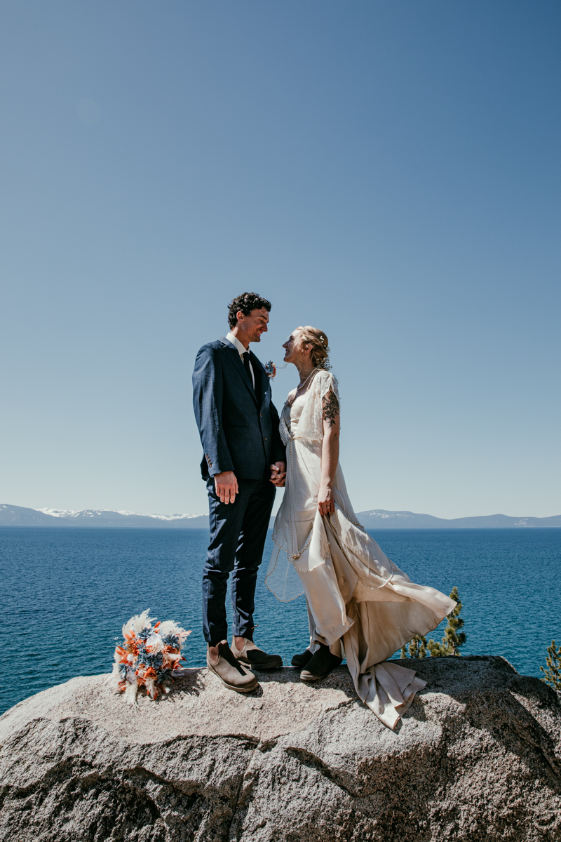 Bride and groom holding hands and smiling at each other on a rock overlooking Lake Tahoe during their Logan Shoals Vista Point elopement