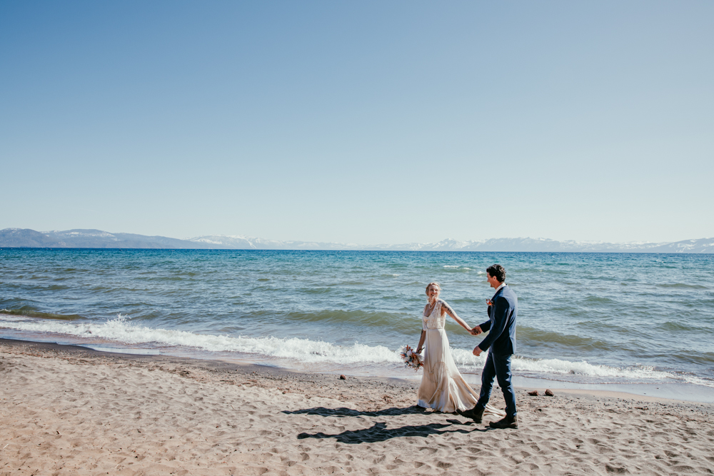 Bride and groom walking hand-in-hand along the shoreline at Kings Beach in Lake Tahoe, with gentle waves and snow-capped mountains in the distance