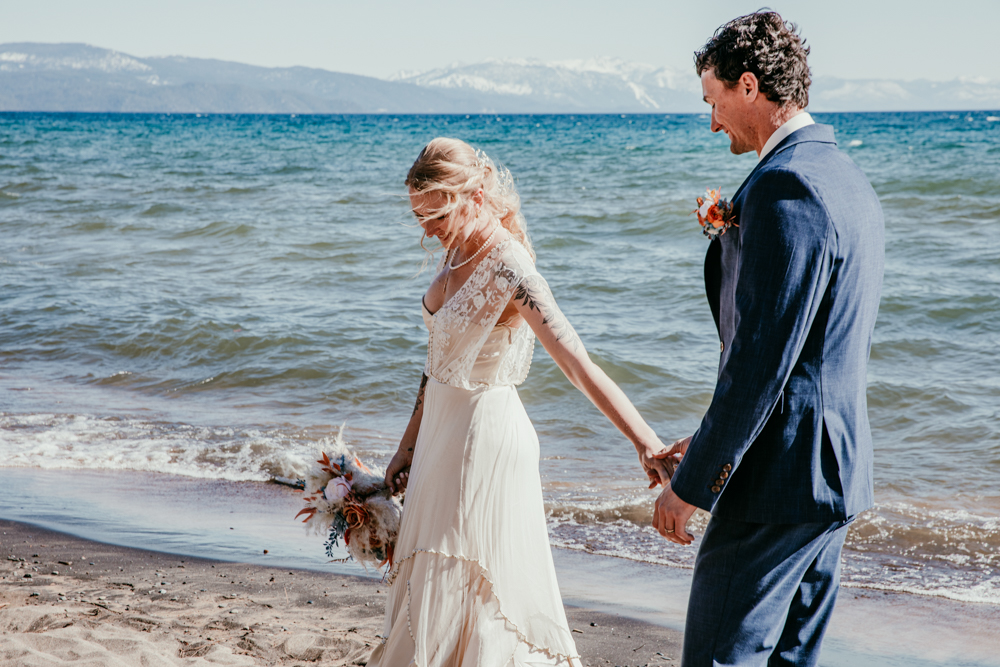 Bride and groom holding hands and walking barefoot along the shore at Kings Beach, with the bride smiling down and Lake Tahoe sparkling behind them