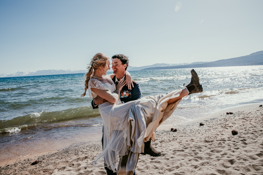 Groom lifting and spinning the bride in his arms on the beach at Kings Beach, both laughing with Lake Tahoe shimmering in the background