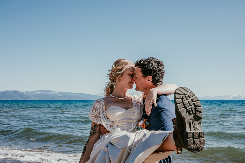 Groom lifts bride on the beach at Kings Beach, Lake Tahoe during their spring elopement, both smiling and soaking in the moment with the lake and mountains in the background.