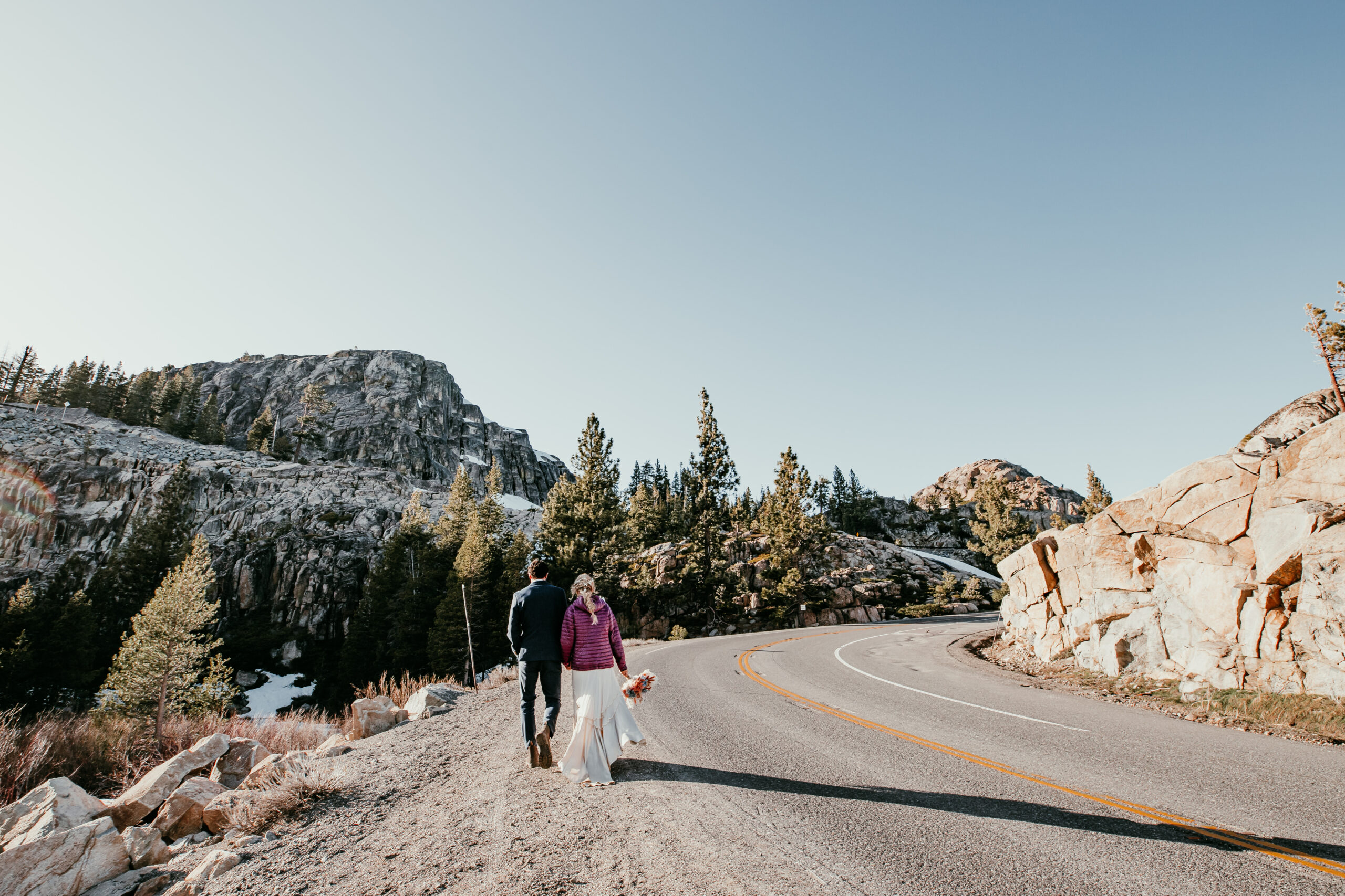 Couple walking along a mountain road surrounded by rocky cliffs and pines during their fall elopement in Lake Tahoe.