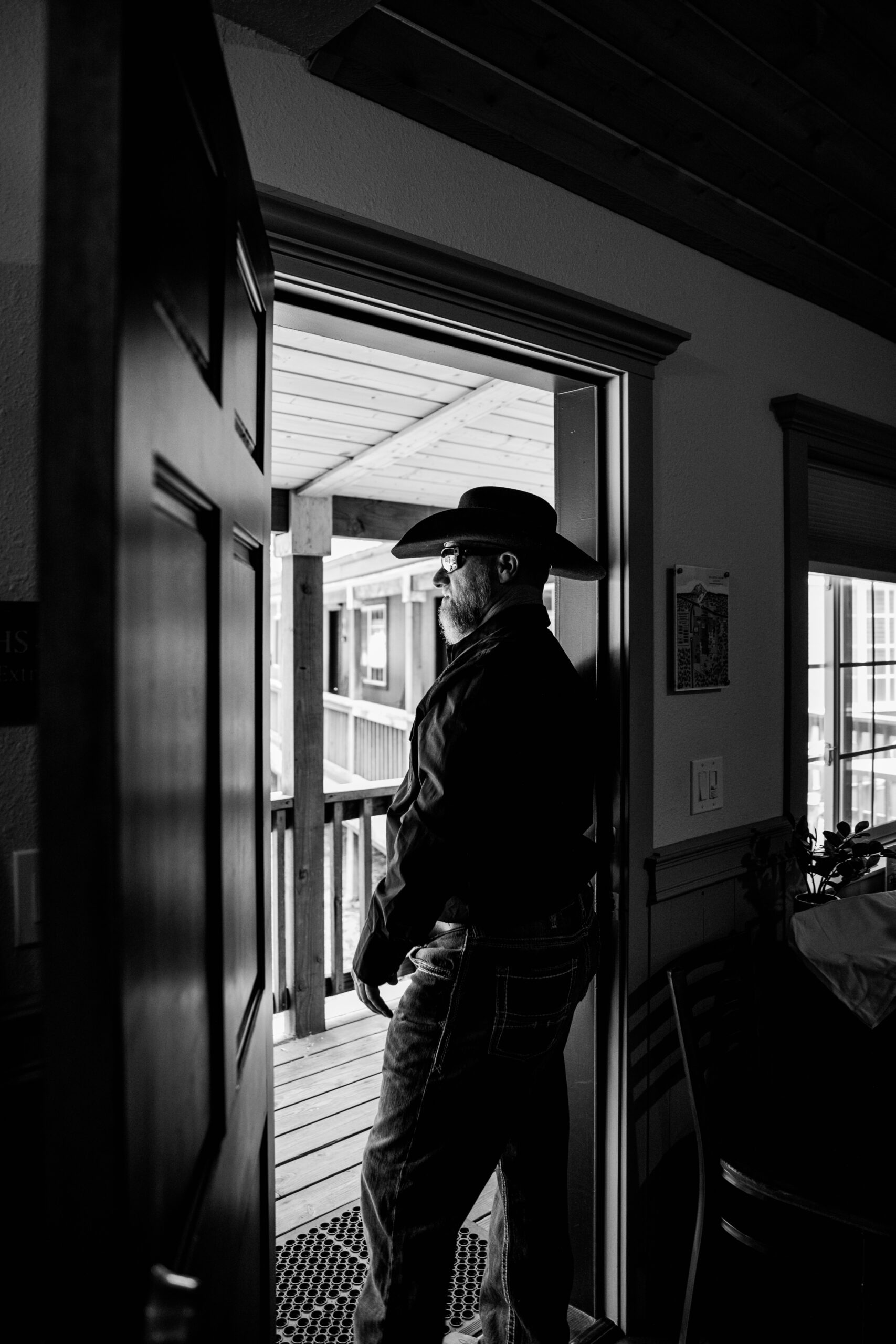 Groom wearing a cowboy hat looking out from a doorway before eloping in Mount Shasta, captured in a black and white candid moment.