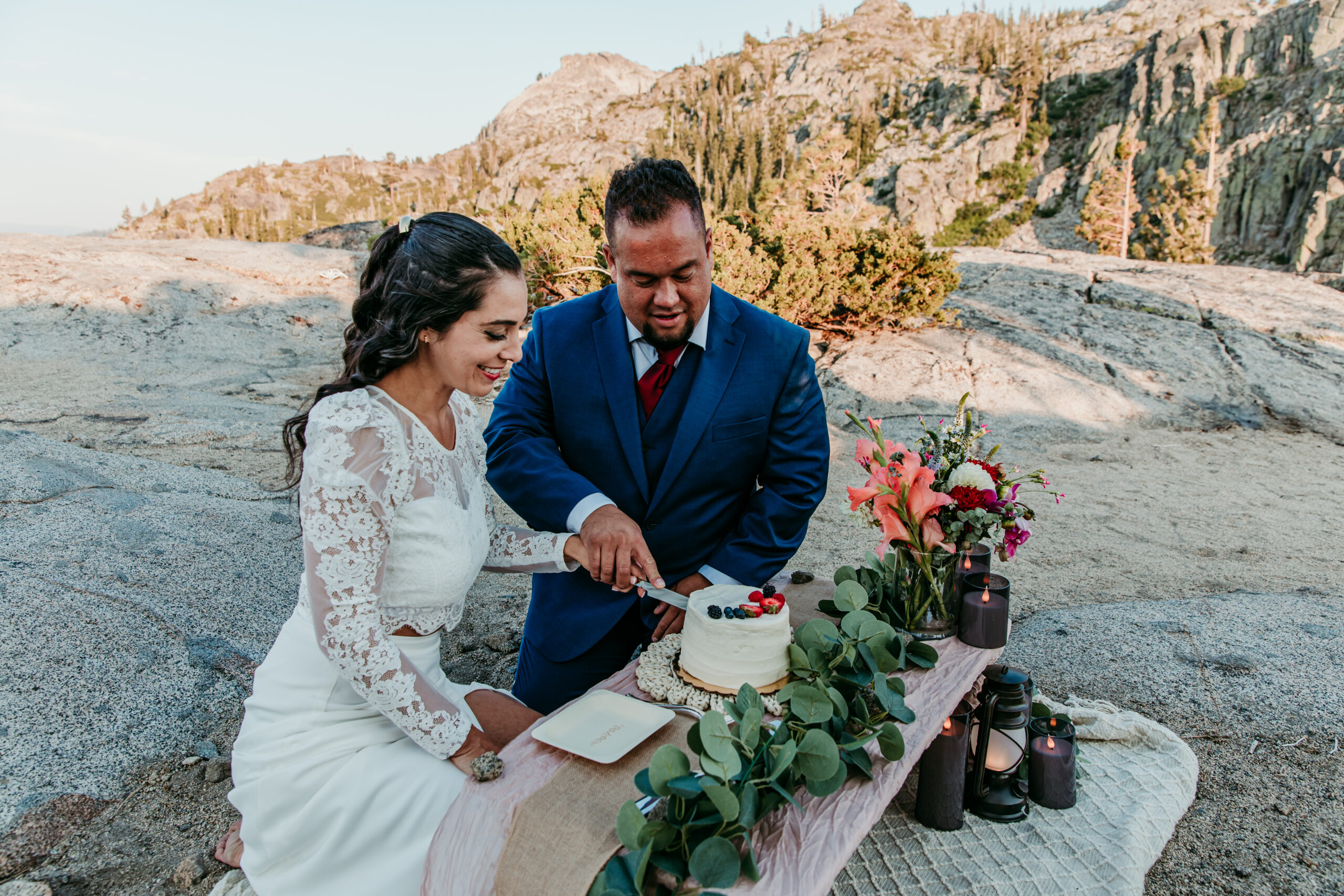 Bride and groom cutting a small cake on a decorated picnic table surrounded by flowers and candles during their elopement in Lake Tahoe.