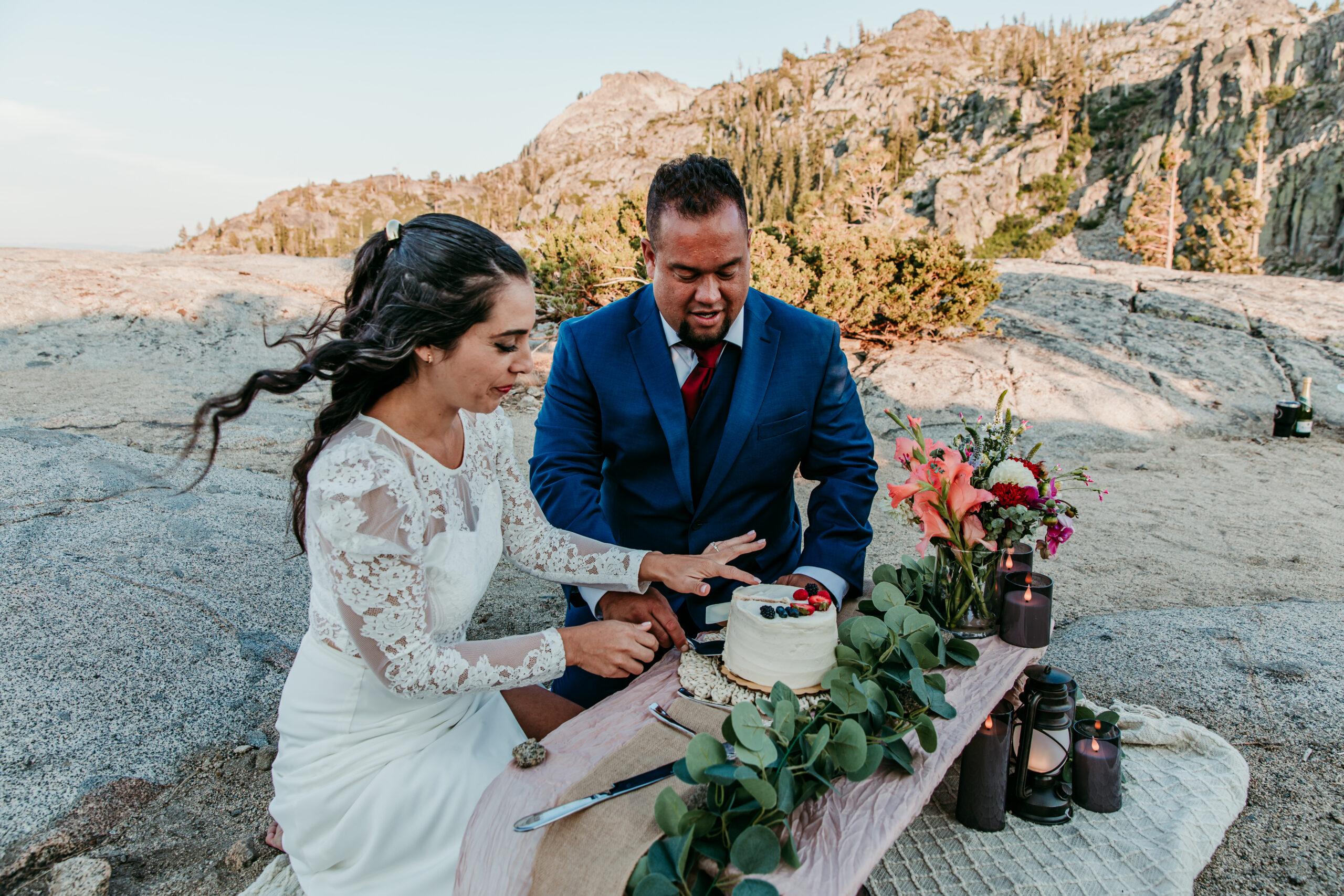 Bride and groom cutting a small cake on a wooden picnic table adorned with flowers and greenery during their elopement in Lake Tahoe.