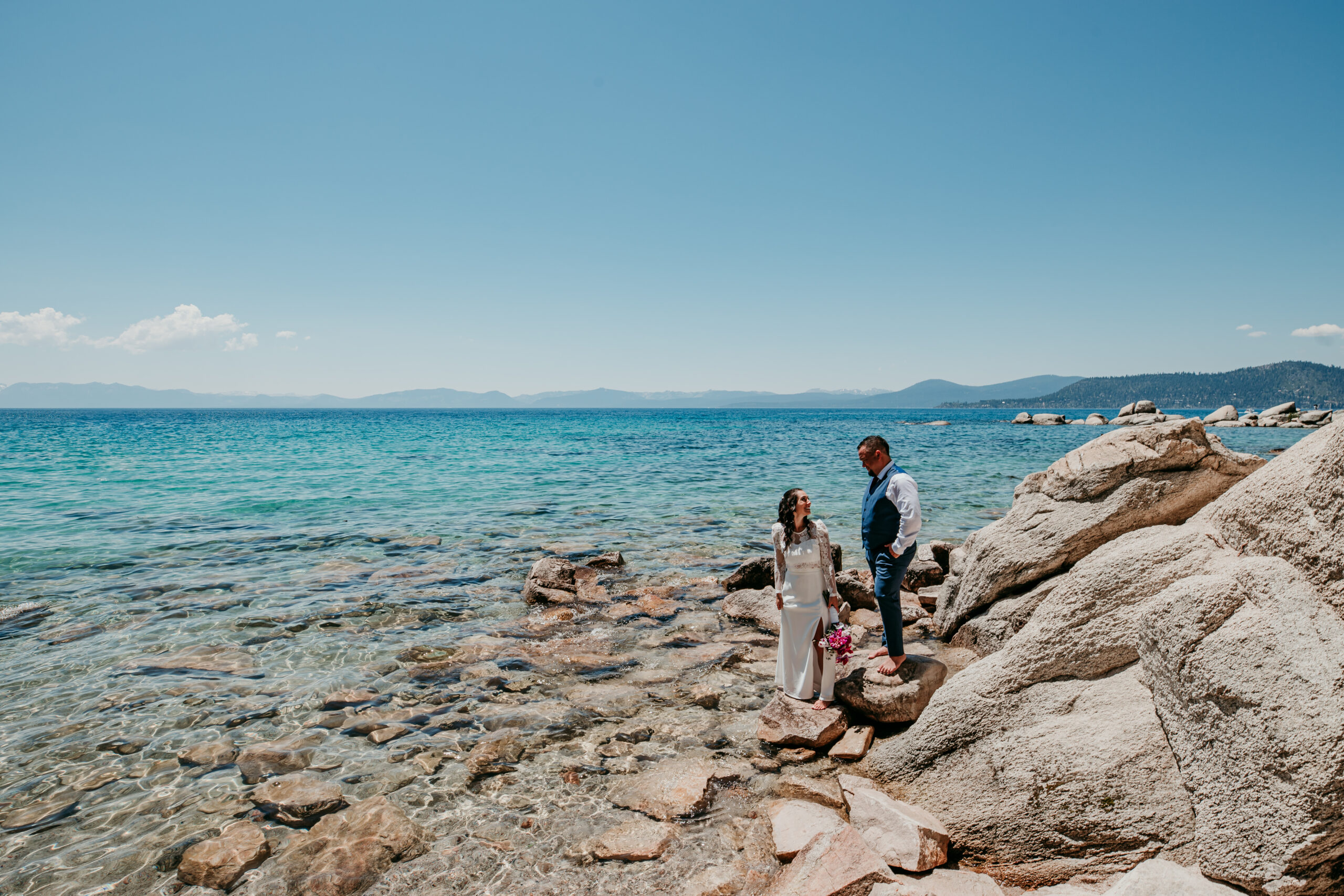 Bride and groom standing on rocky shoreline during their elopement in Lake Tahoe with turquoise water in the background.