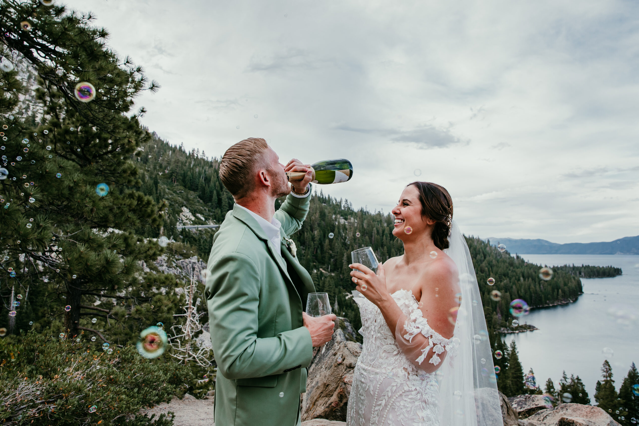 Bride laughing while groom drinks champagne straight from the bottle during their Emerald Bay elopement celebration overlooking Lake Tahoe.